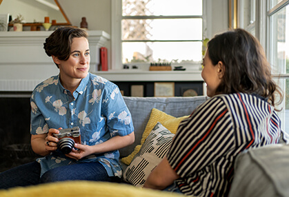 Woman with an instant-film camera, sitting on a couch and discussing LYBALVI® (olanzapine and samidorphan) Care Support with another woman