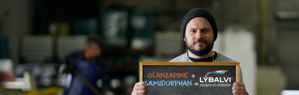 Learn about the effects of LYBALVI® on symptoms Man standing in a warehouse, holding a sign that shows the components of LYBALVI® (olanzapine and samidorphan)